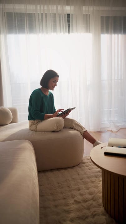 Woman using a tablet while sitting cross-legged on a sofa in morning sunlight