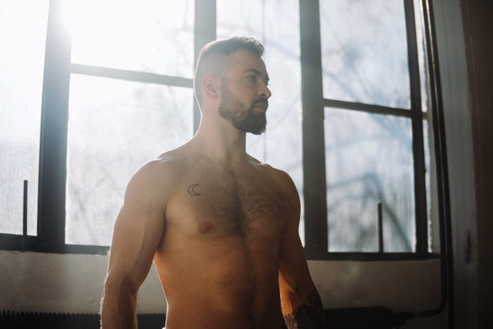 Shirtless Man Standing Pensive In Sunlit Yoga Studio By Large Window