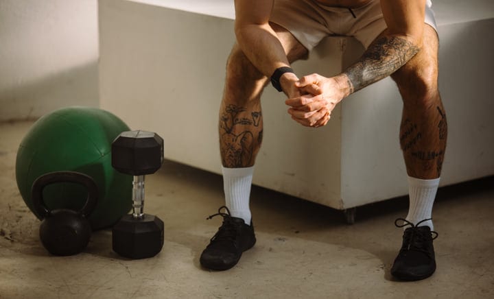 Tattooed Man Sits On A Bench Next To Weights In Gym