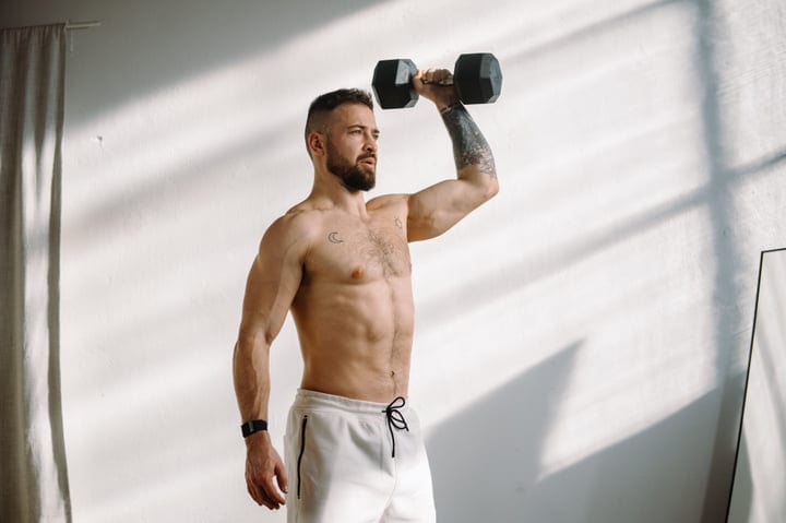 Determined Man With Tattoos Lifting A Dumbbell Overhead During His Indoor Workout
