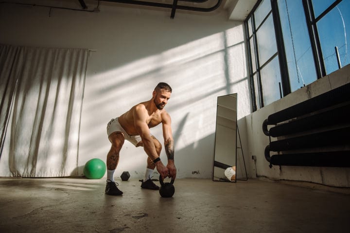Man With Tattoos Squatting To Lift Kettlebell In Modern Gym