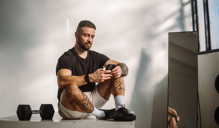 Bearded Man With Tattoos Sitting On A Bench Using His Smartphone In A Gym