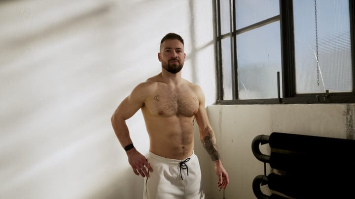 Muscular Man With Tattoos Standing Shirtless In A Minimalist Indoor Gym Space